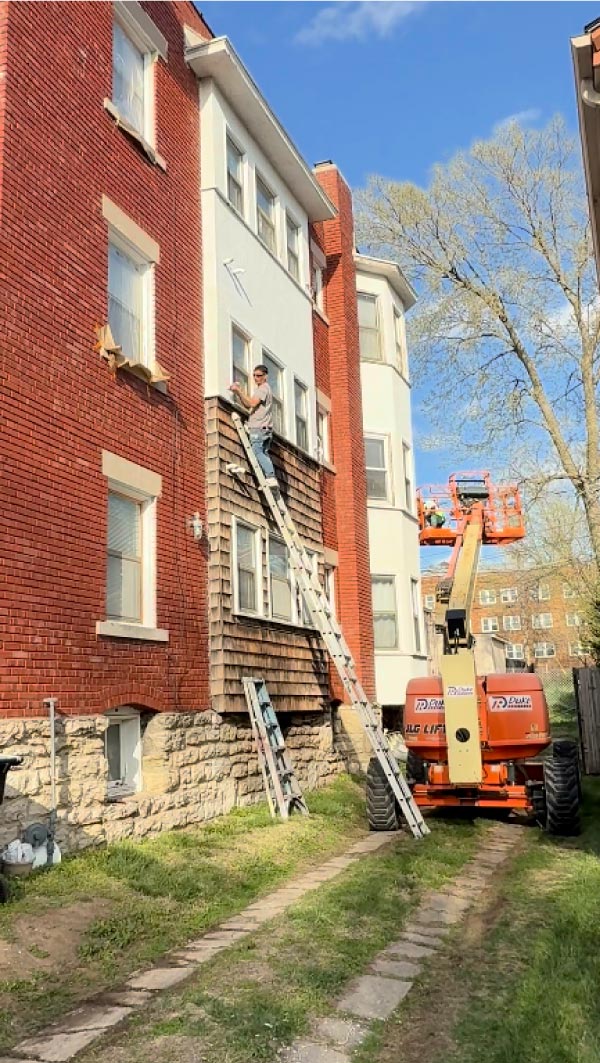 professional painters from Elite77 Painters LLC using a high-reach boom lift and extension ladders to perform exterior painting and maintenance on a multi-story red brick and tan residential building in Kansas City.