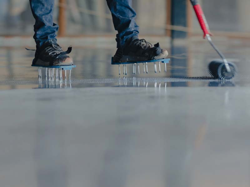 A professional installer wearing spiked shoes to walk across a wet surface while using a roller to apply a smooth, high-gloss epoxy flooring coating